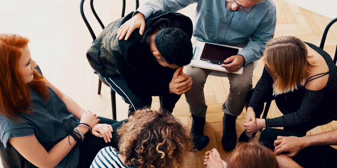 drug addicts sitting in a circle and one man with his head in his hands and the counselor consoling him